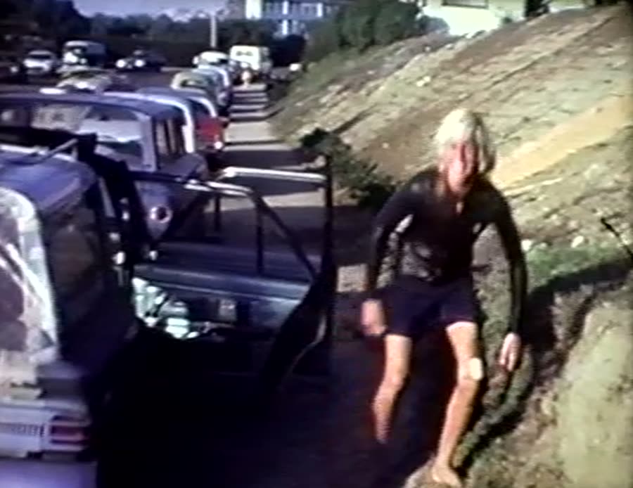 A blond surfer in a wetsuit climbing up from the beach between parked cars